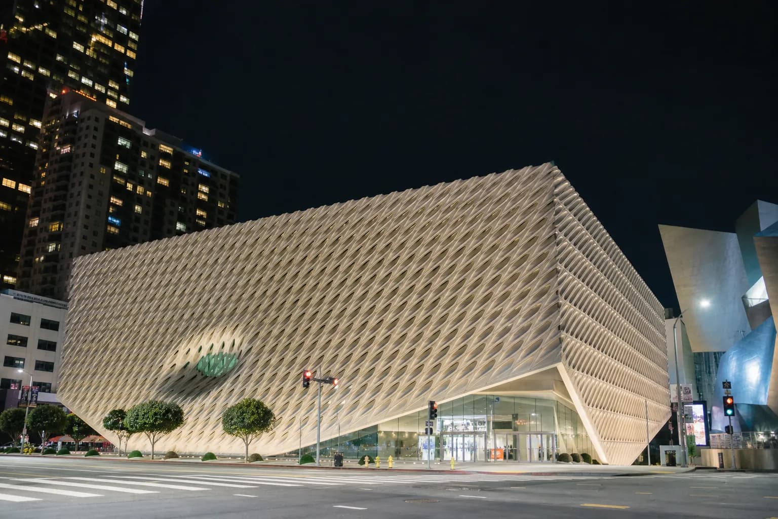 The exterior of a modern Los Angeles art gallery illuminated at night, featuring clean concrete lines and large glass windows glowing with warm interior light.