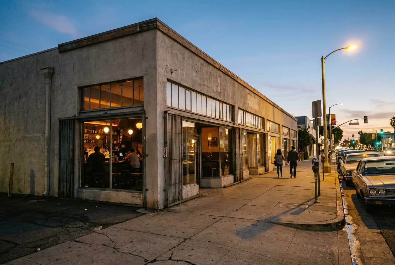 Exterior view of a low-lit wine bar on Hollywood Boulevard in Thai Town at dusk, warm light visible through large windows, cars parked along the street.