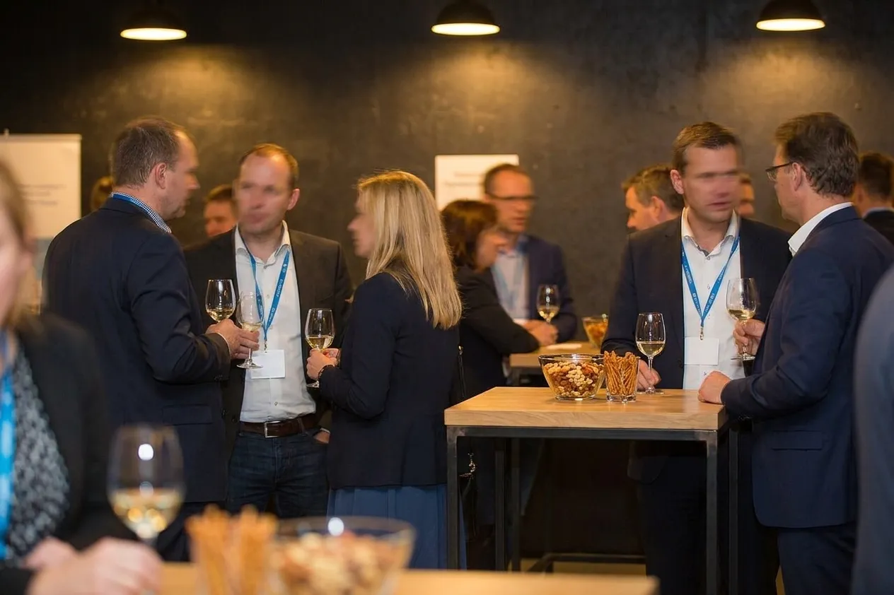Small standing groups at an evening industry mixer with glasses and name-badge lanyards beside a high table holding a bowl of snacks