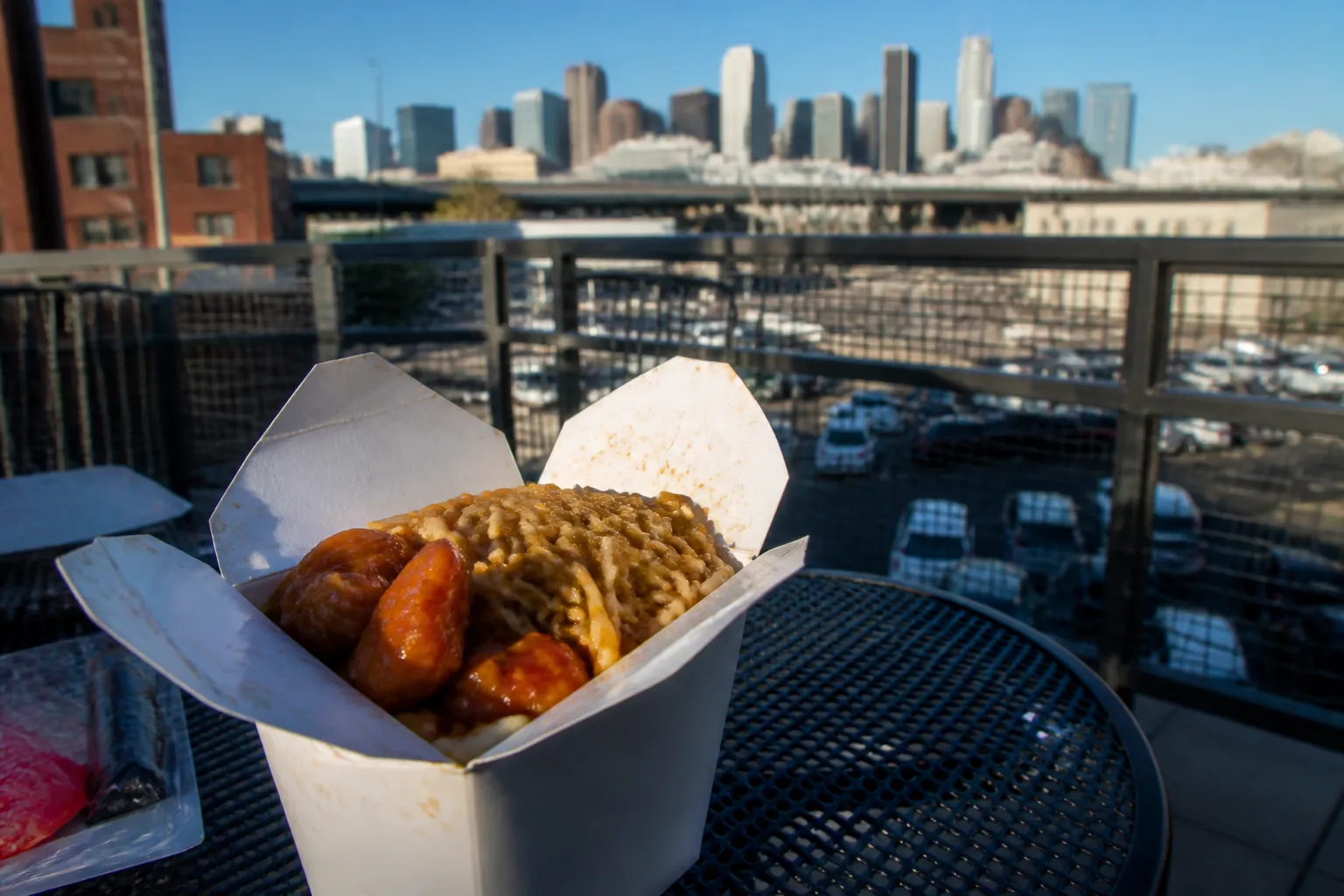 Close-up of orange chicken and chow mein in a takeout box with a blurred Downtown LA skyline