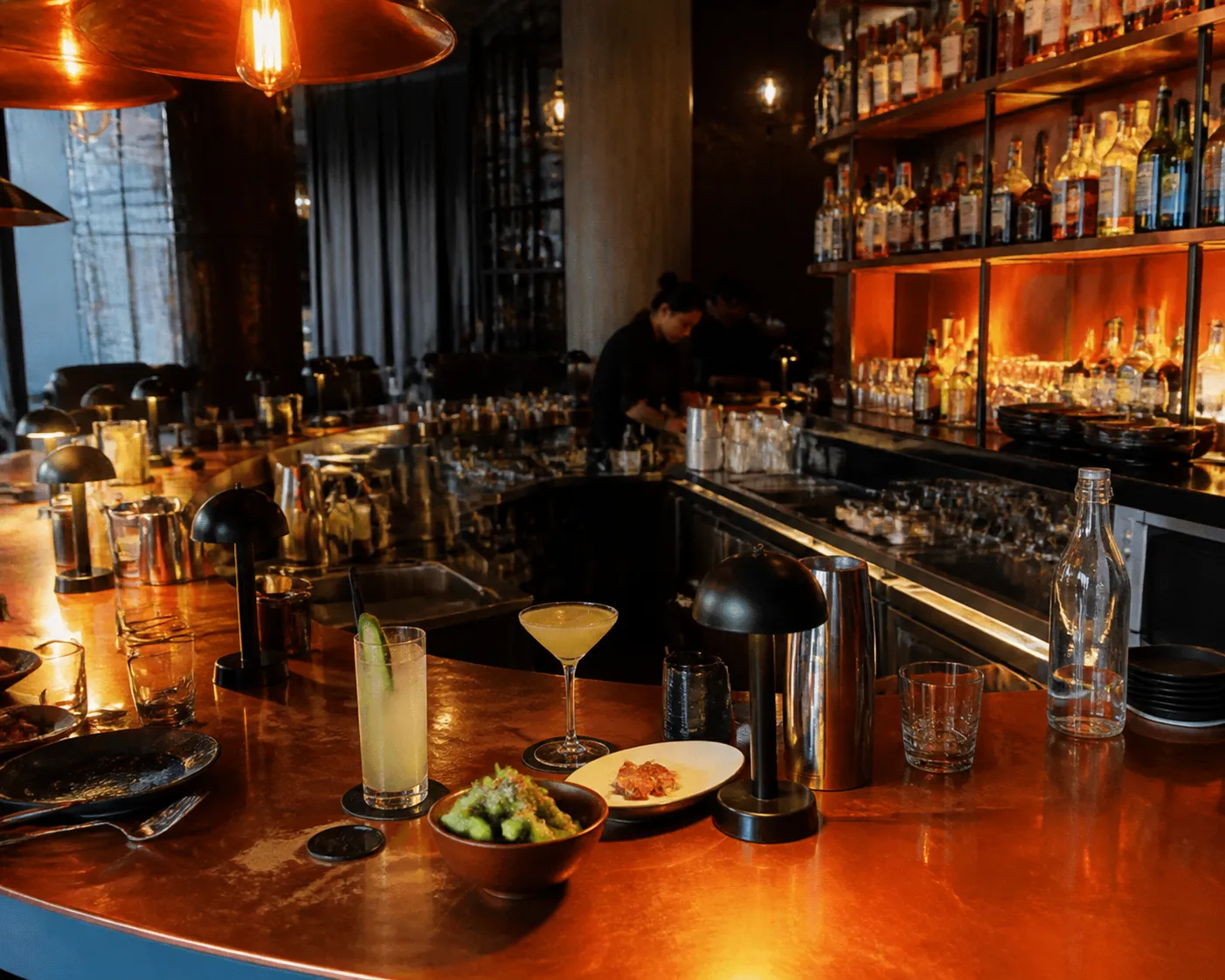 A close-up of a modern copper bar counter in a DTLA restaurant with cocktails and food plates.