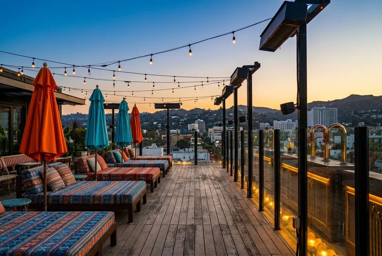 Hollywood rooftop terrace at dusk with patterned daybeds, tropical umbrellas and string lights overlooking Hollywood Hills skyline under amber sky.