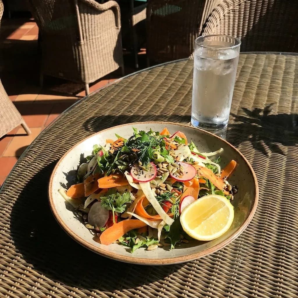 A large salad plate on an outdoor table in direct sun with shaved vegetables, herbs, seeds and a wedge of citrus beside a glass of iced water