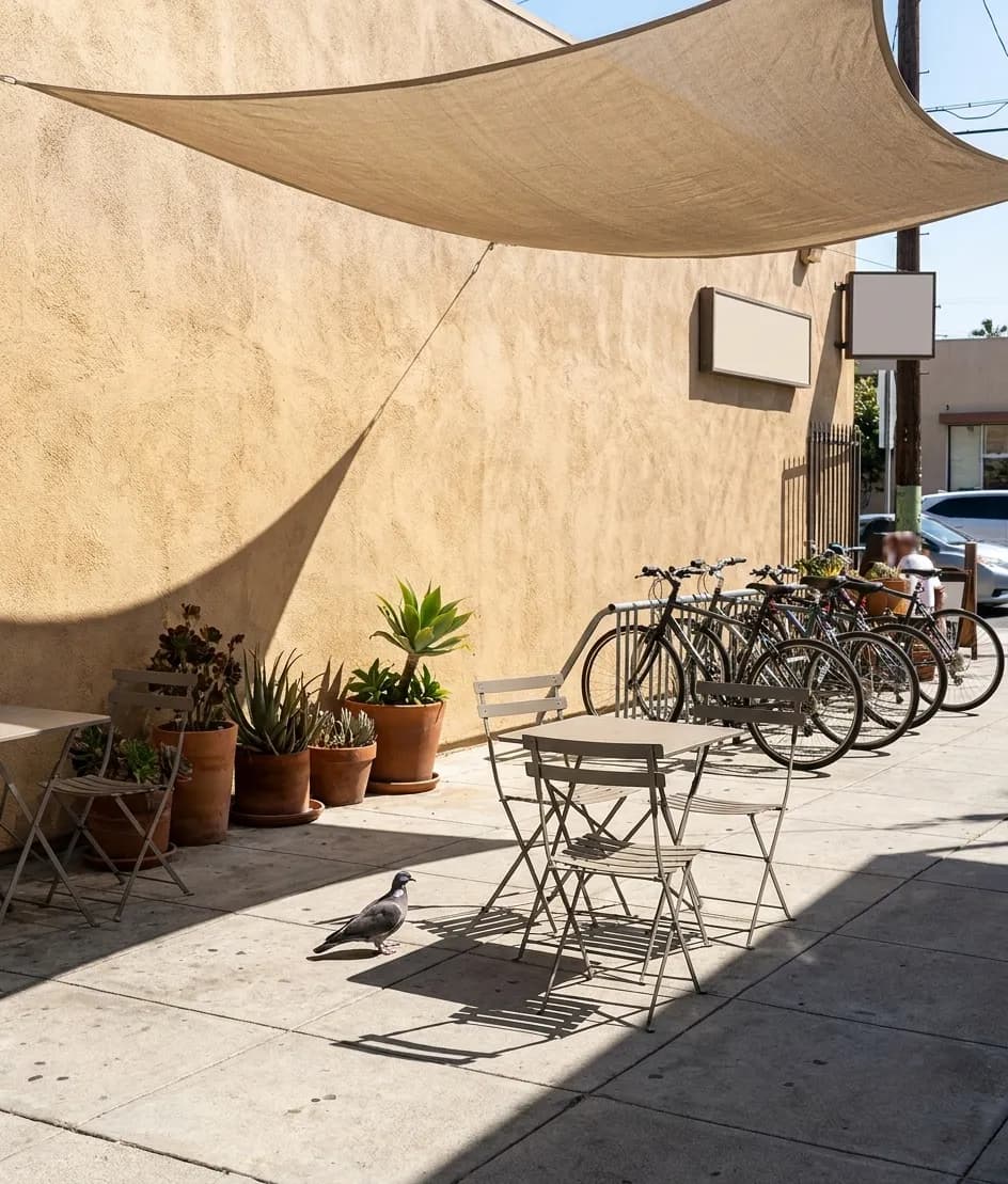 A Los Angeles cafe patio at midday with small metal tables under a shade sail, potted succulents and bicycles at a rack in bright sun