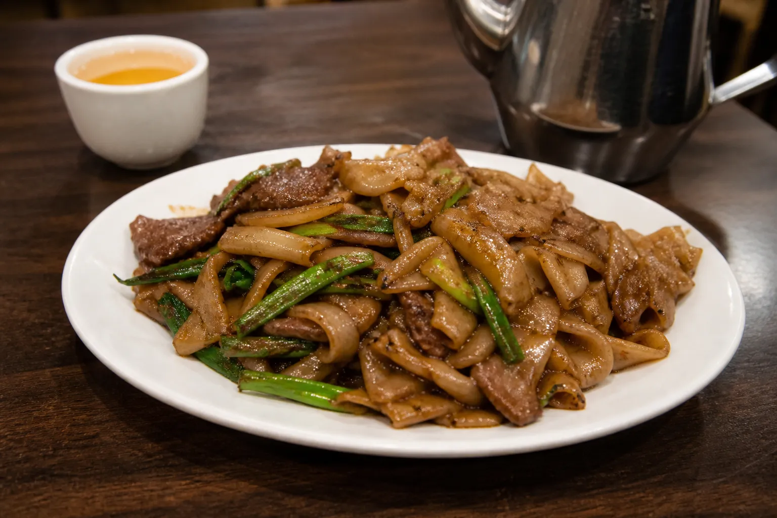 A steaming plate of dry-fried beef chow fun glistening with savory oils on a dark laminate tabletop, flanked by wooden chopsticks and a small dish of house-made chili crisp.