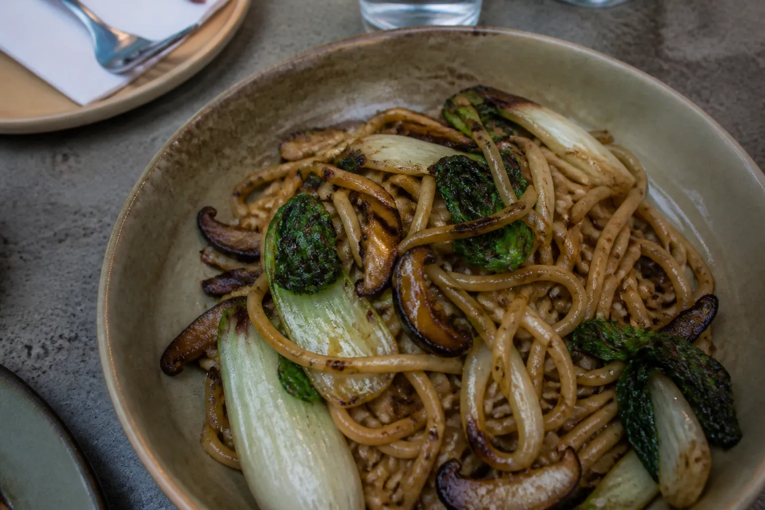 A bowl of thick hand-pulled noodles with charred greens on a concrete counter.