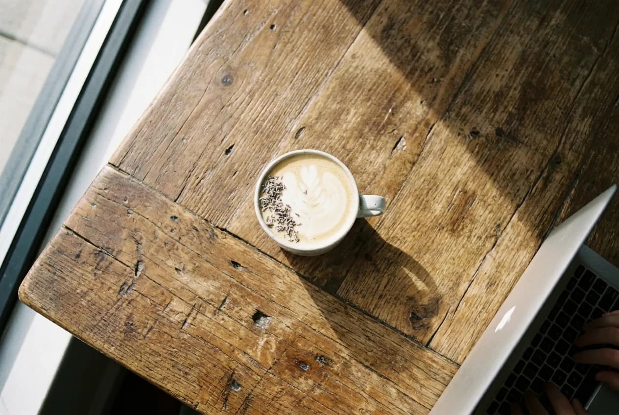 Overhead view of honey lavender latte in ceramic mug on wooden communal table with laptop keyboard partially visible at edge of frame