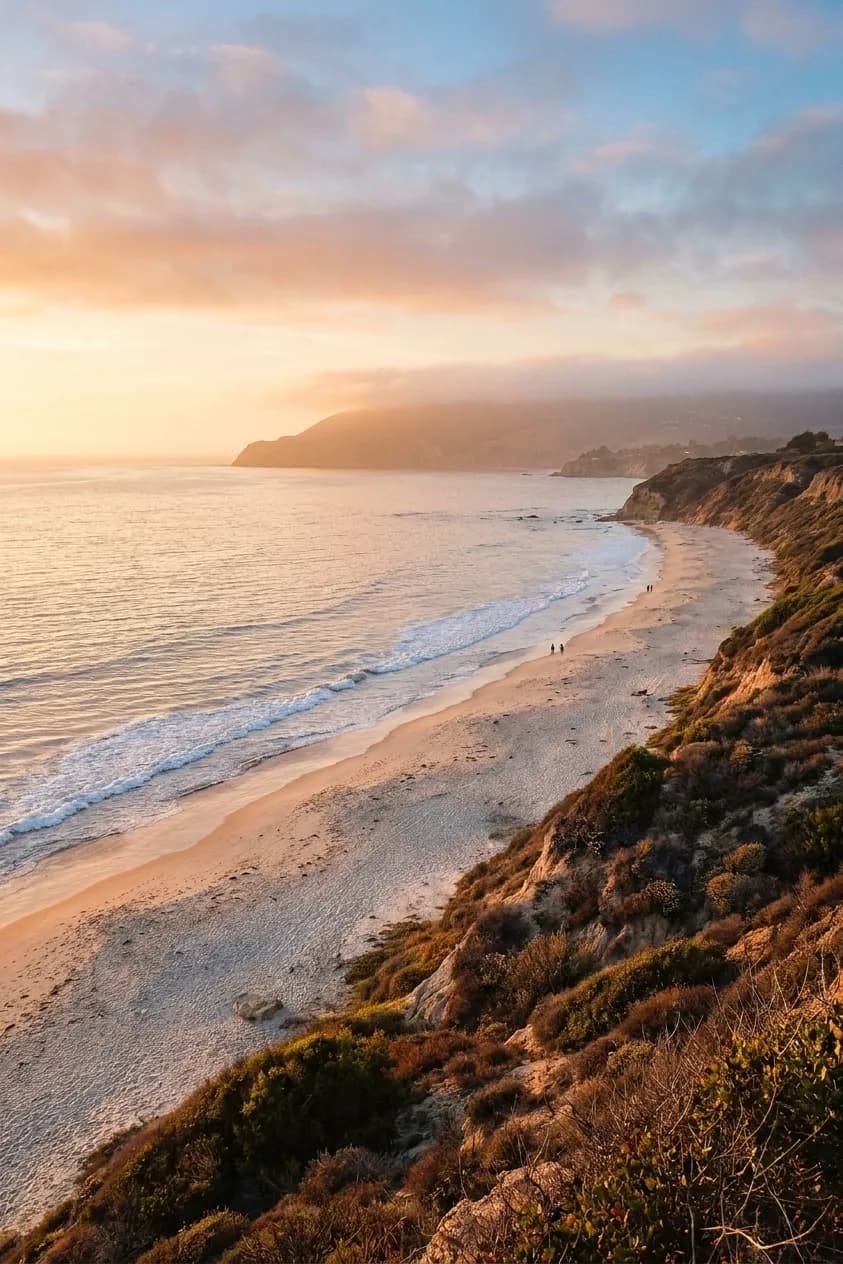 A wide Southern California shoreline in late afternoon, with pale sand curving away beneath dry chaparral bluffs, small breaking waves and a headland softened by marine haze in the distance
