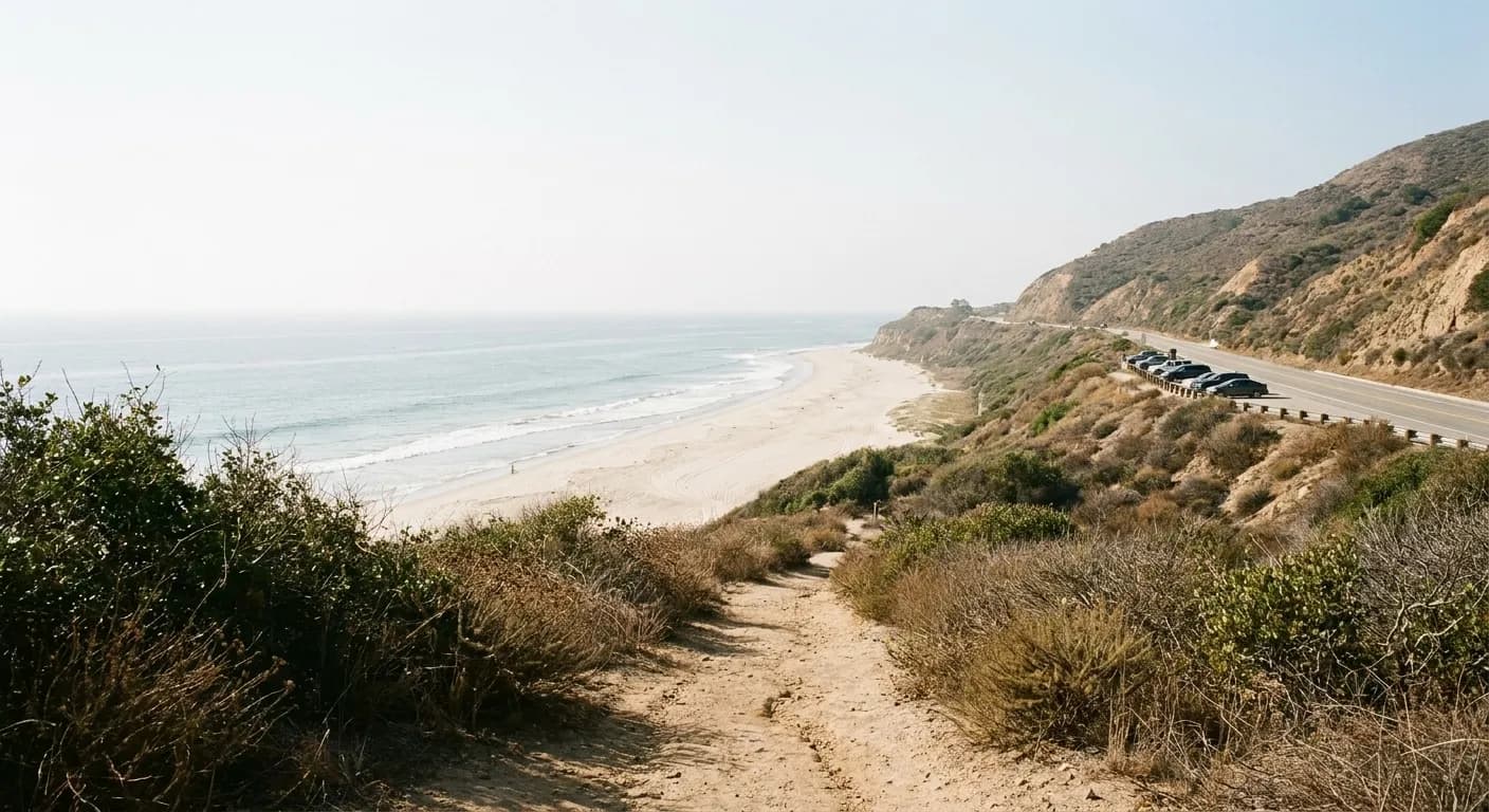 A Southern California beach seen from the bluff above with a coast highway cut into the hillside and cars parked on the shoulder