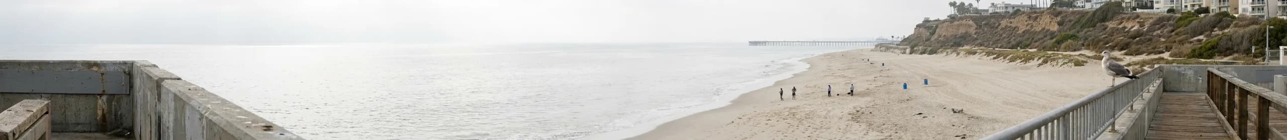 A quiet weekday stretch of Los Angeles County coast, wide pale sand curving away below dry scrub bluffs with a distant pier on the horizon