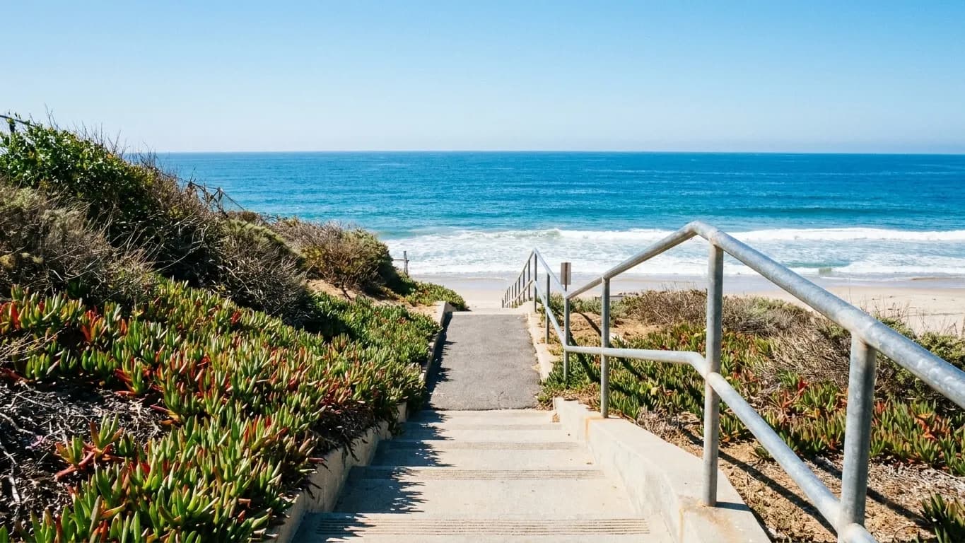 A narrow public access path and concrete steps dropping between ice-plant banks toward a Los Angeles beach, ocean bright at the end