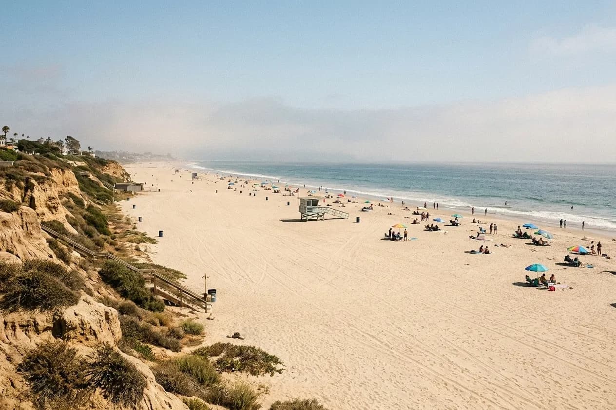 A broad pale Los Angeles beach in the afternoon with scattered umbrellas, a stilted lifeguard tower and a bluff along one side
