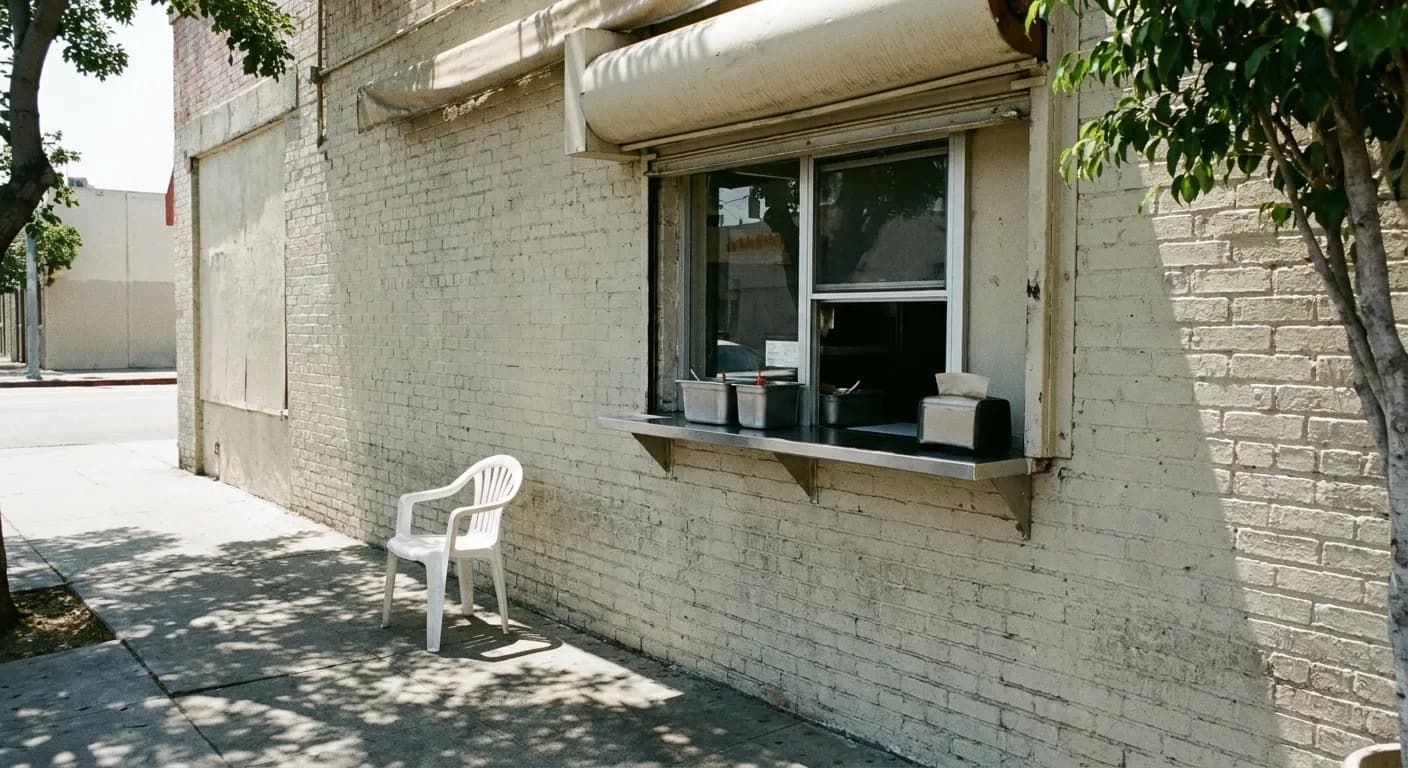 A walk-up food window in a painted brick wall on a Los Angeles corner, with a steel shelf of condiments and dappled light on the sidewalk