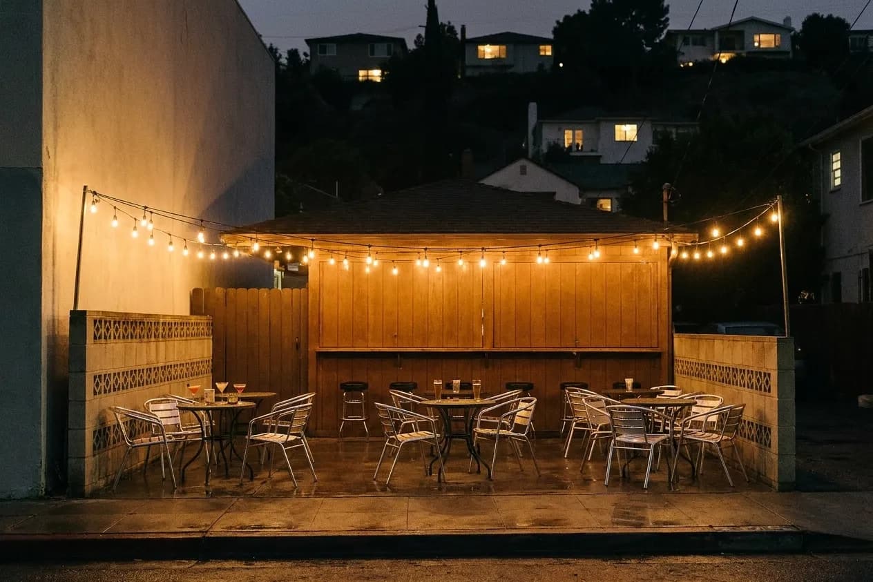 A small Los Angeles bar patio after sunset with metal tables behind a breeze-block wall and a strand of bulbs overhead