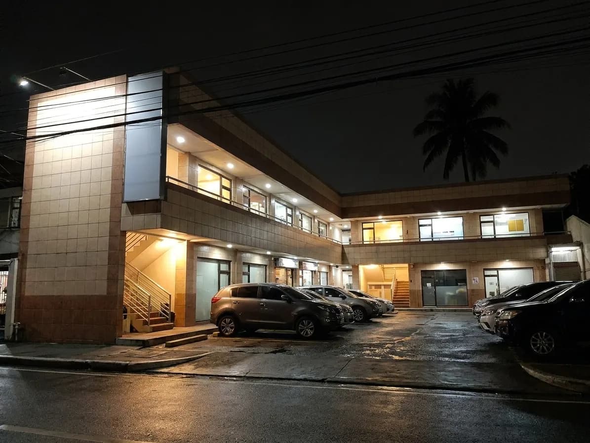 A two-storey Koreatown mini-mall after dark with lit stairwells to upper-floor units and cars nosed into the small forecourt