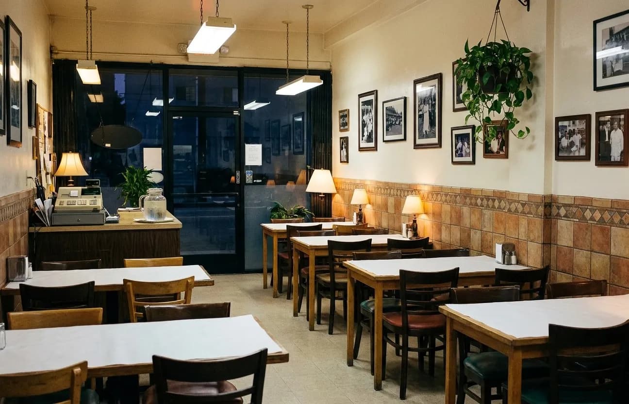A small family-run Los Angeles restaurant in the evening with eight paper-covered tables and mismatched chairs, a tiled dado, framed photographs and a service counter at the back