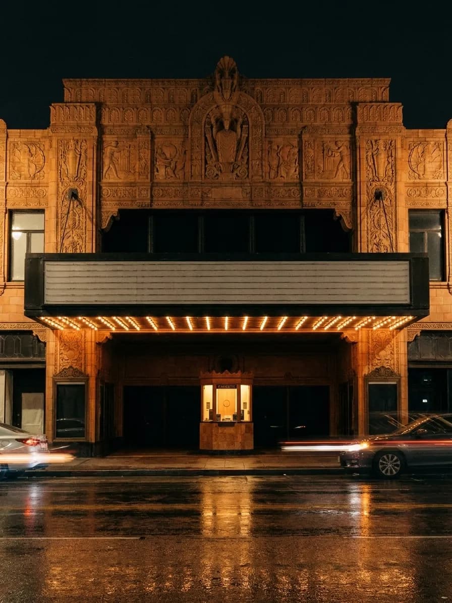The ornate terracotta frontage of a 1920s Los Angeles theatre at night, a blank bulb-studded marquee above a warmly lit recessed entrance