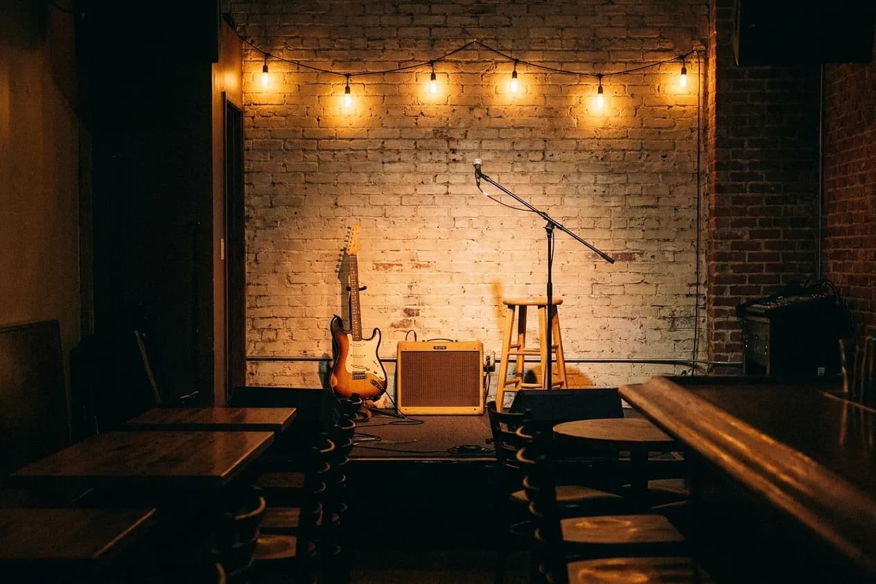 A guitar, small amp and mic stand on a tiny corner stage in a Los Angeles bar, warm bulbs strung above a painted brick wall