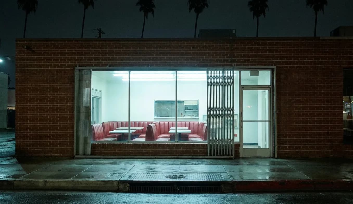 A Los Angeles restaurant frontage at midnight, its wide window glowing white against a dark street with empty booths inside