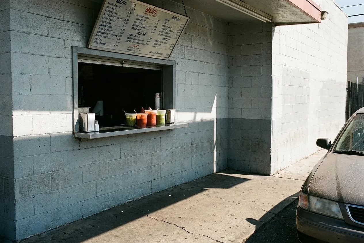 A walk-up food counter cut into a painted cinderblock wall in Los Angeles, with a shallow condiment ledge and hard midday sun on the sidewalk