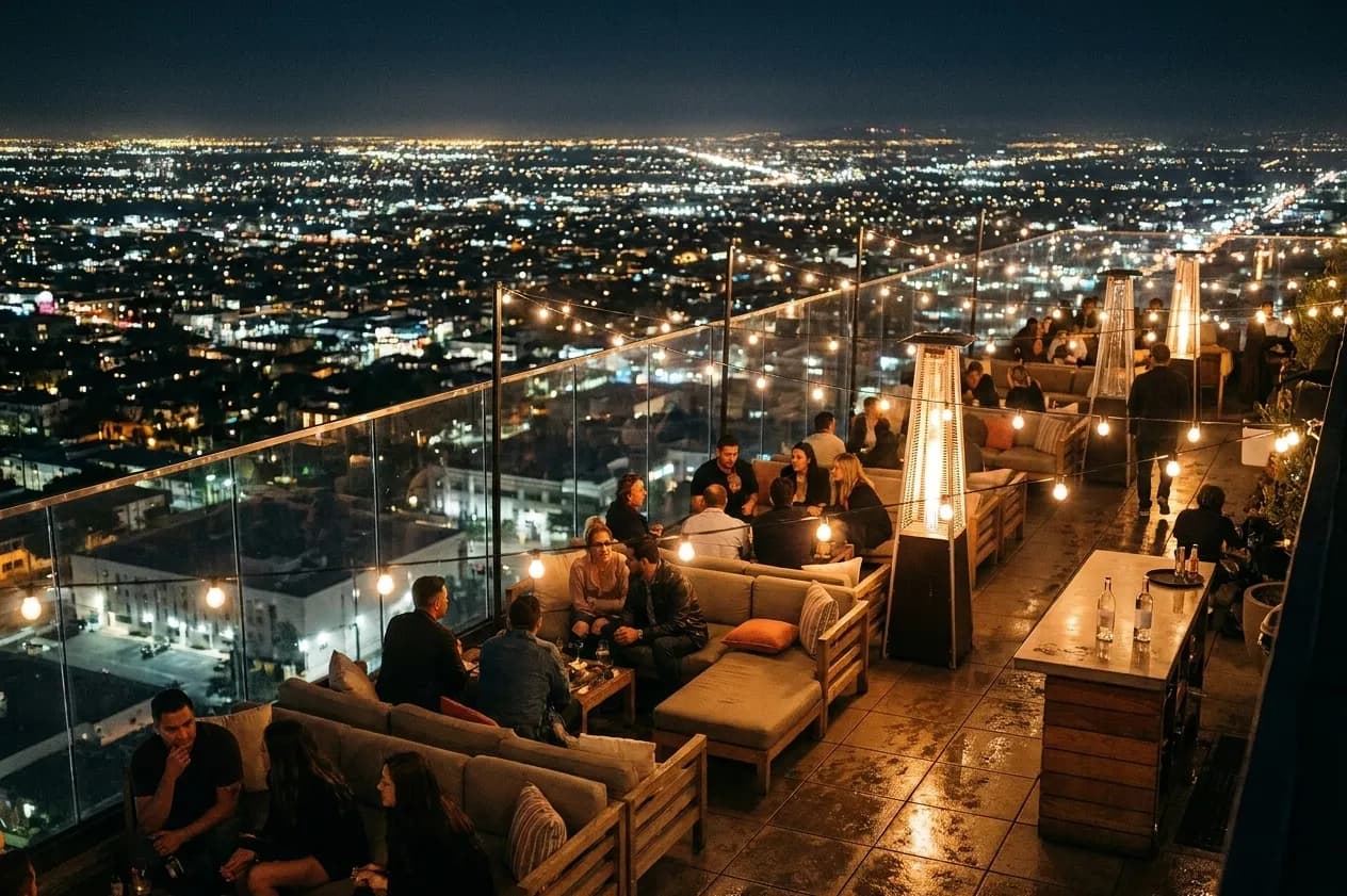 A rooftop bar terrace in Los Angeles after dark with string lights, low couches and the city grid glowing below