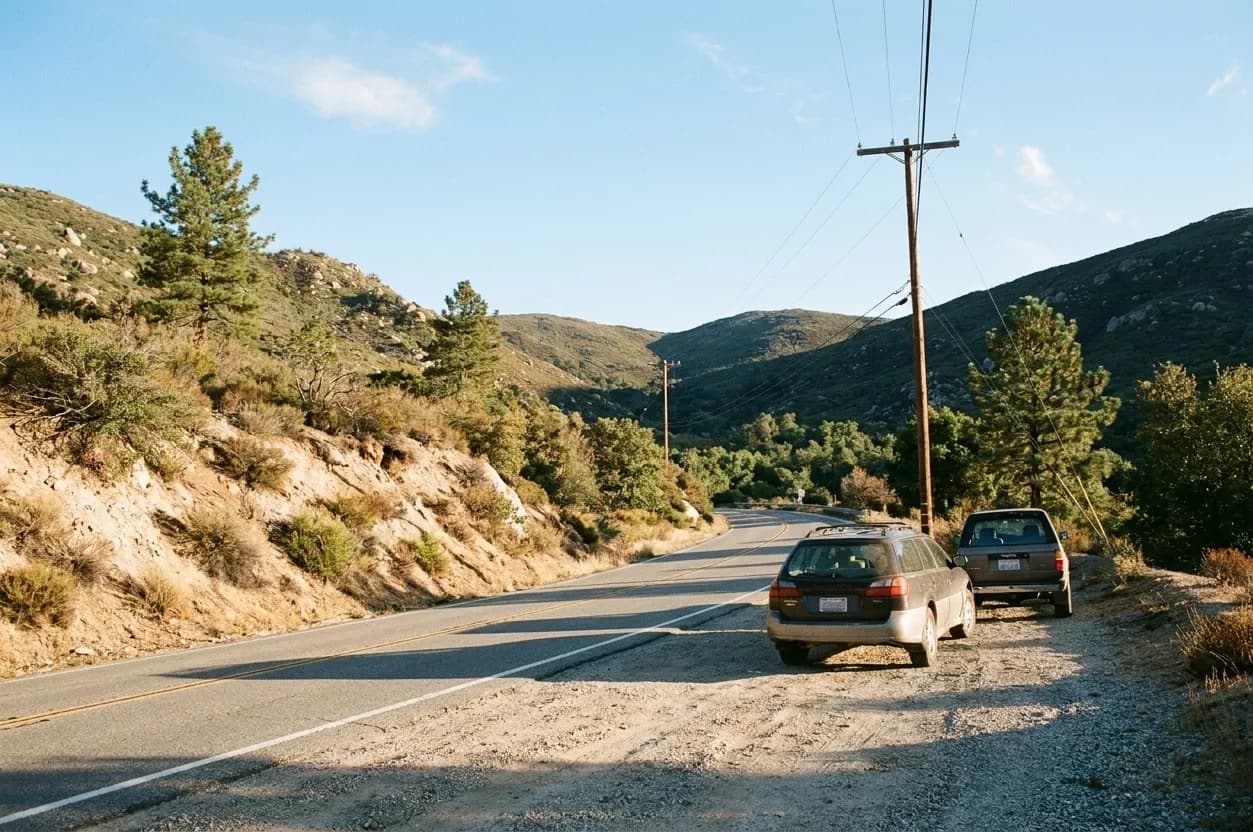 A two-lane mountain road curving through chaparral at sunrise with a gravel pullout holding two parked cars