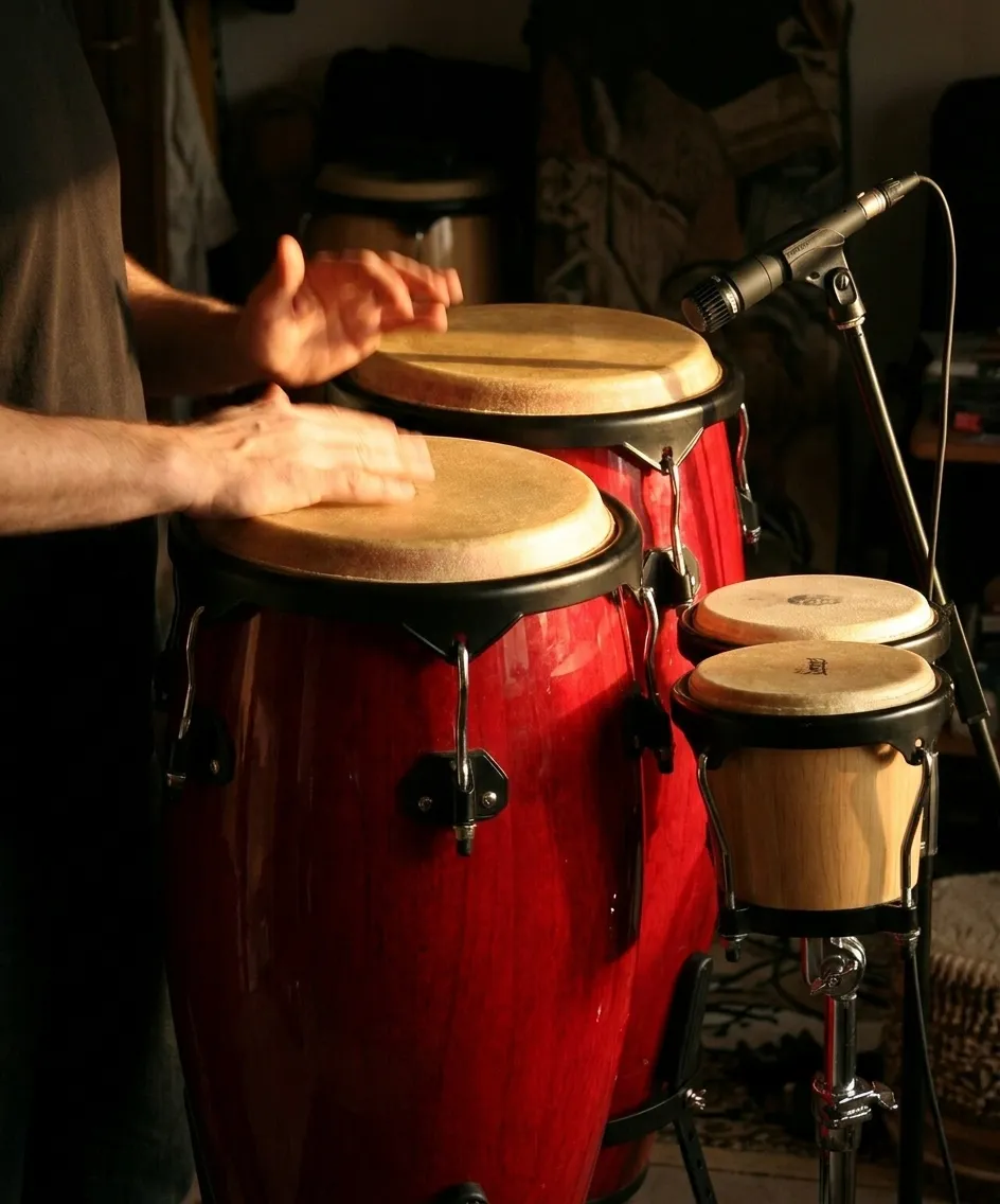 A pair of congas and a set of bongos on stands beside a microphone with a player's hands in motion under warm side light