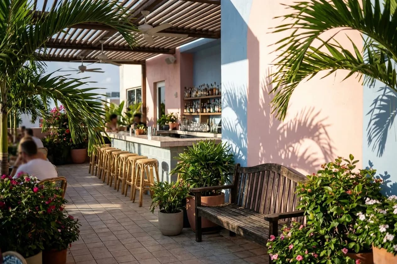 A Miami open-air bar terrace in the late afternoon with rattan stools at a tiled bar and palm fronds throwing hard shadows on a pastel wall