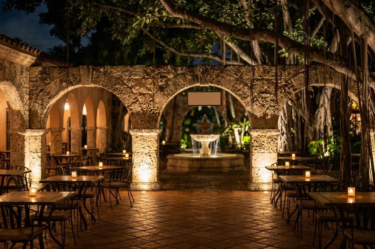 A Coral Gables courtyard at night with coral-stone arches, a tiled floor and small candlelit tables under banyan branches