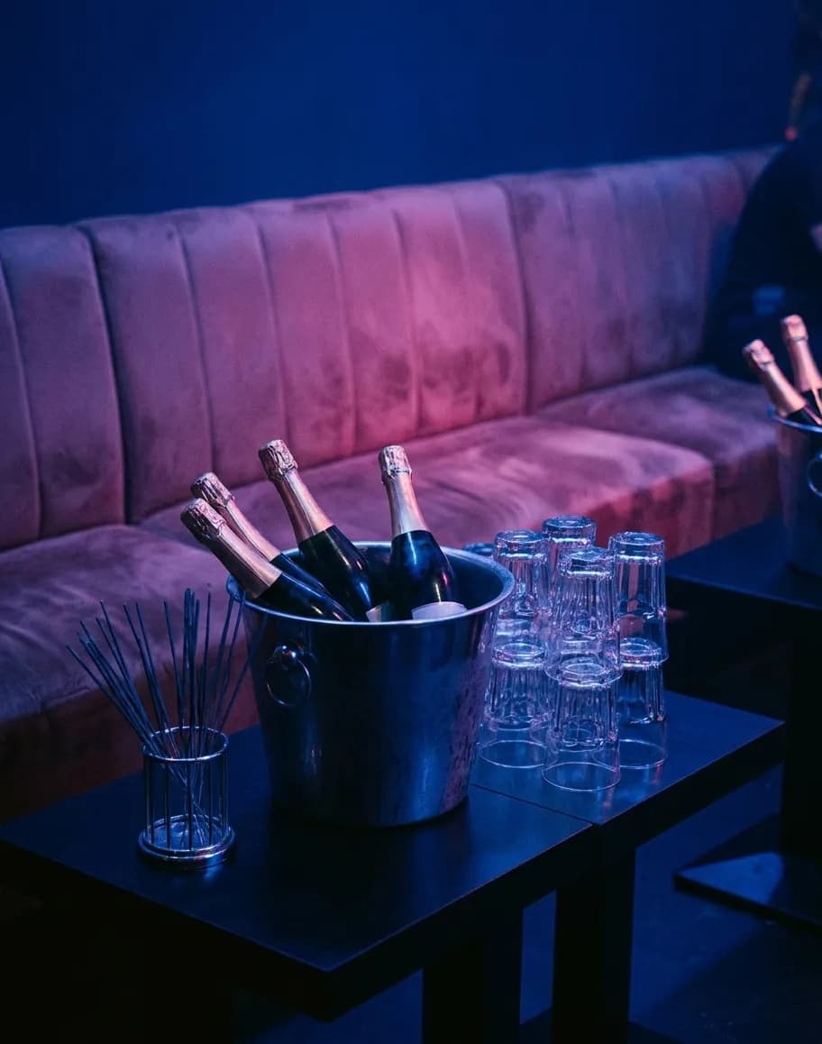 A club table set for a group with an ice bucket holding bottles, a stack of glasses and a low banquette behind under blue and pink lighting