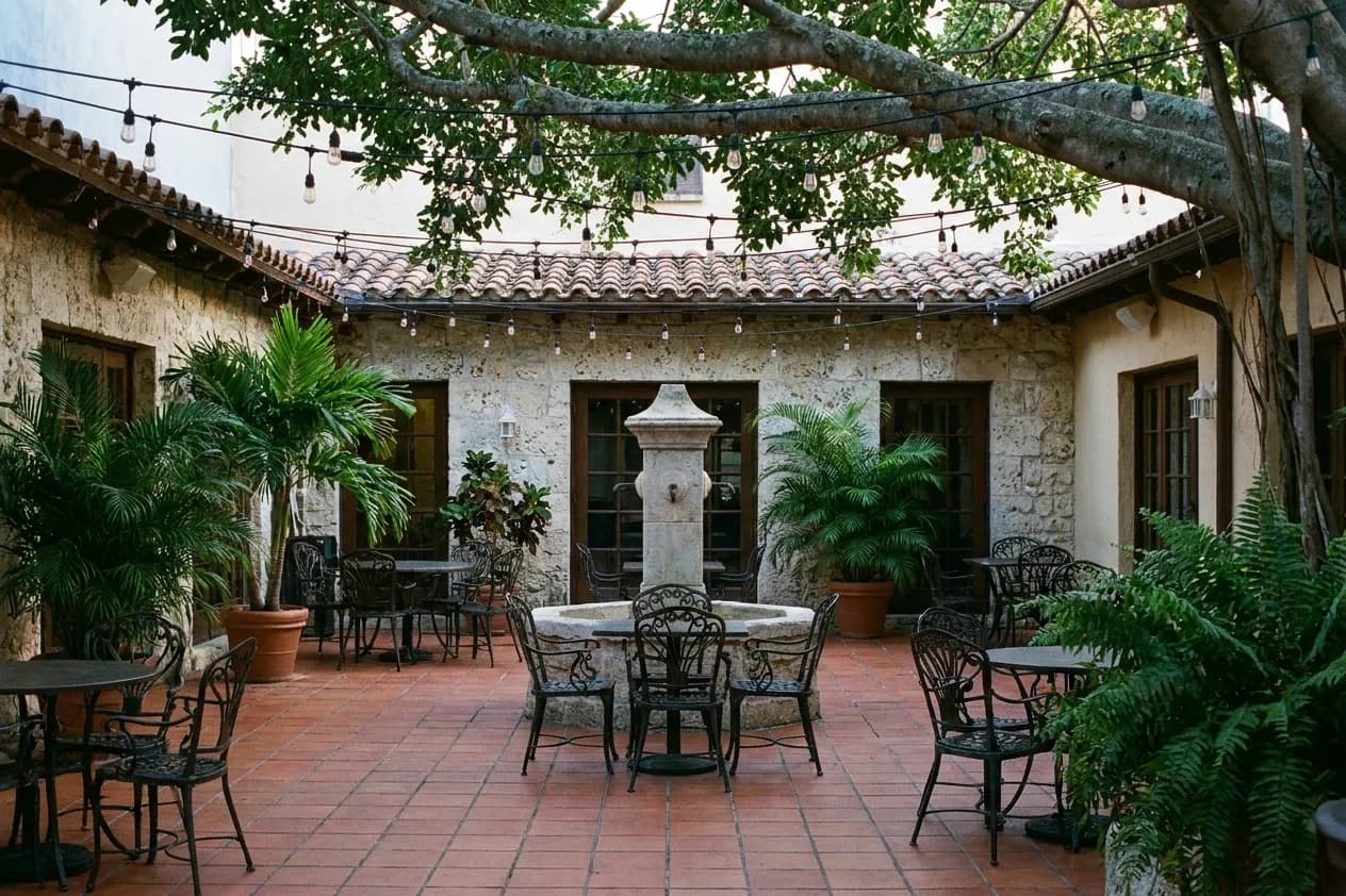 A Coral Gables restaurant courtyard at blue hour with terracotta tiles, wrought-iron chairs, a stone fountain and banyan branches overhead