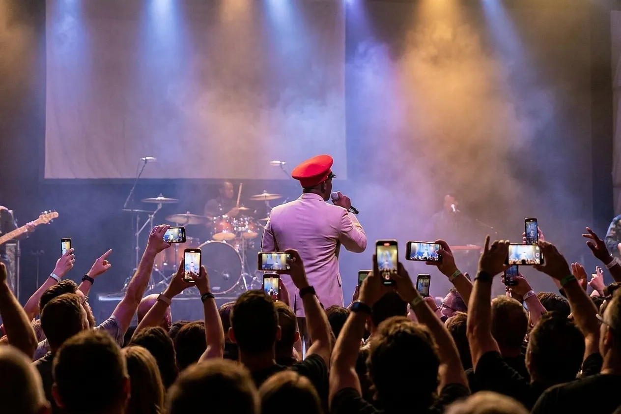 A band mid-set on a low stage seen from behind the crowd with raised phones, coloured wash lights and a blank backdrop behind the drums