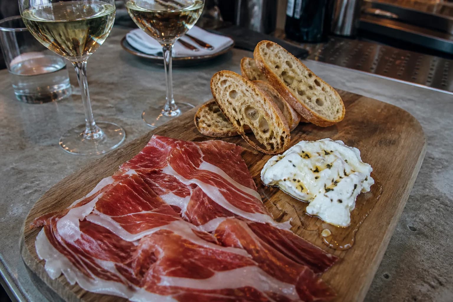 A wooden serving board topped with Spanish cured meats, cheese, bread, and glasses of white wine on a modern concrete bar.