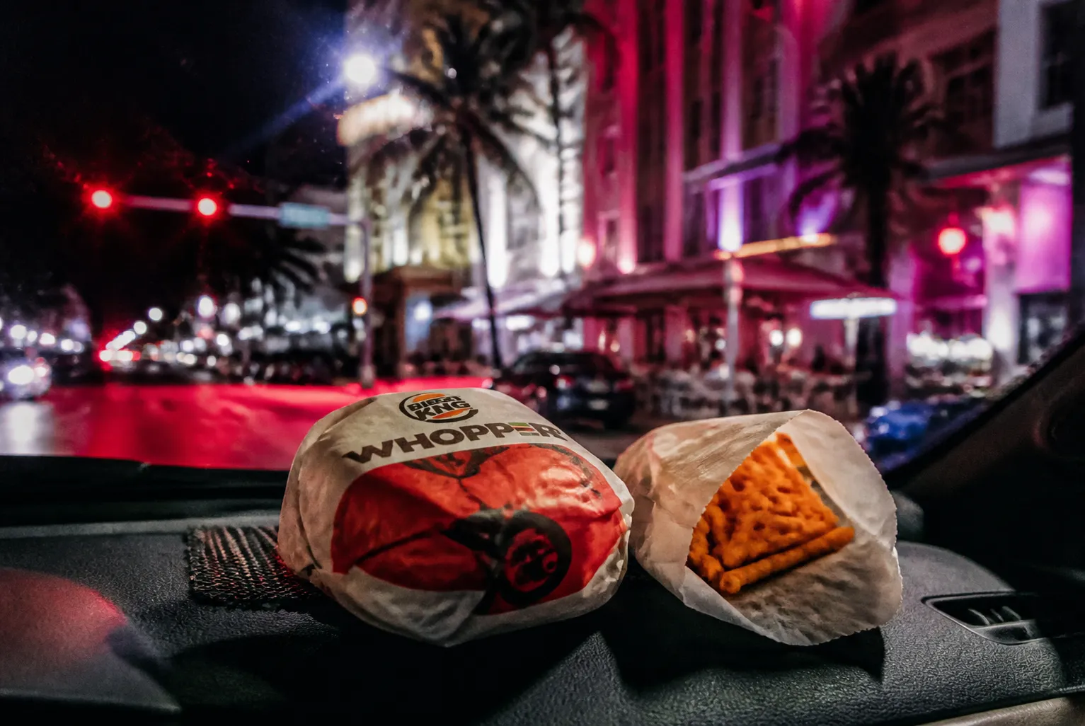 A yellow taxi driving past neon-lit Art Deco buildings on a palm-lined South Beach street at night.