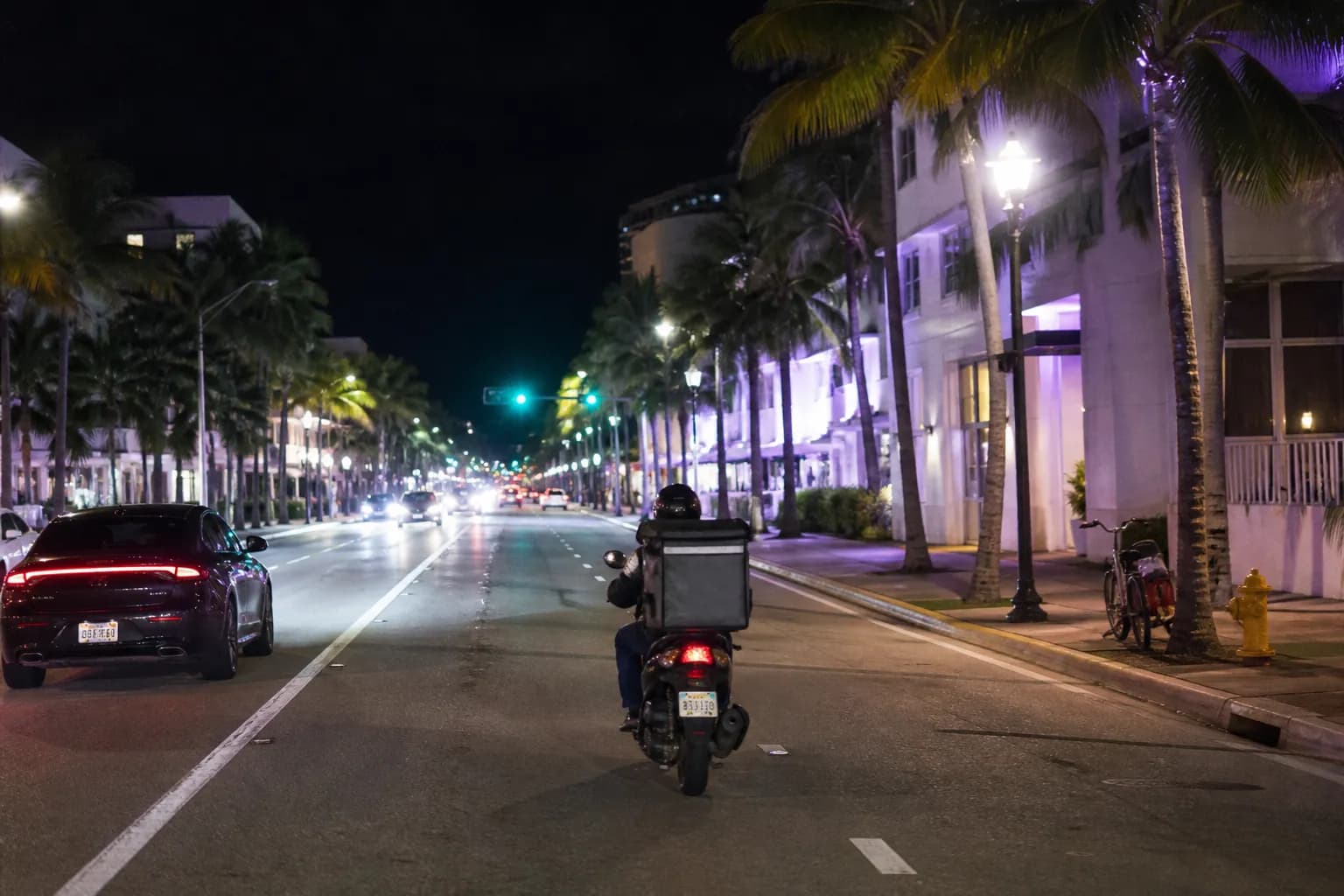 A delivery courier riding a scooter at night past palm trees in South Beach Miami.