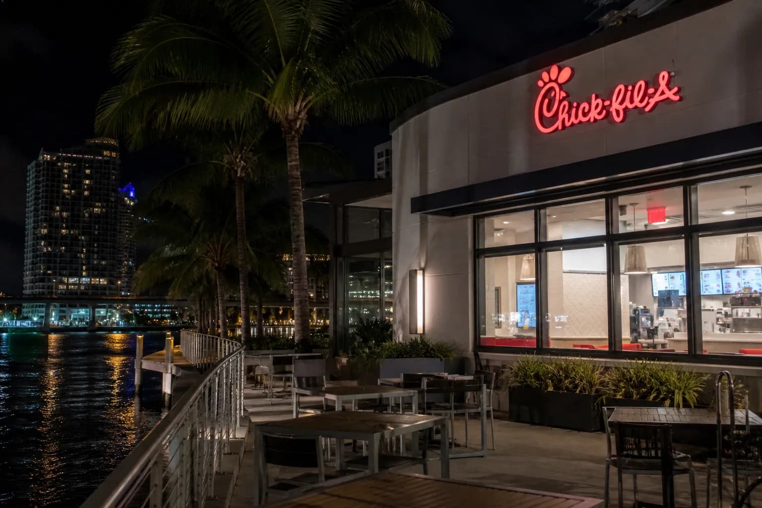 A modern Chick-fil-A drive-thru exterior illuminated at night with palm trees in the background under a dark blue Miami sky.