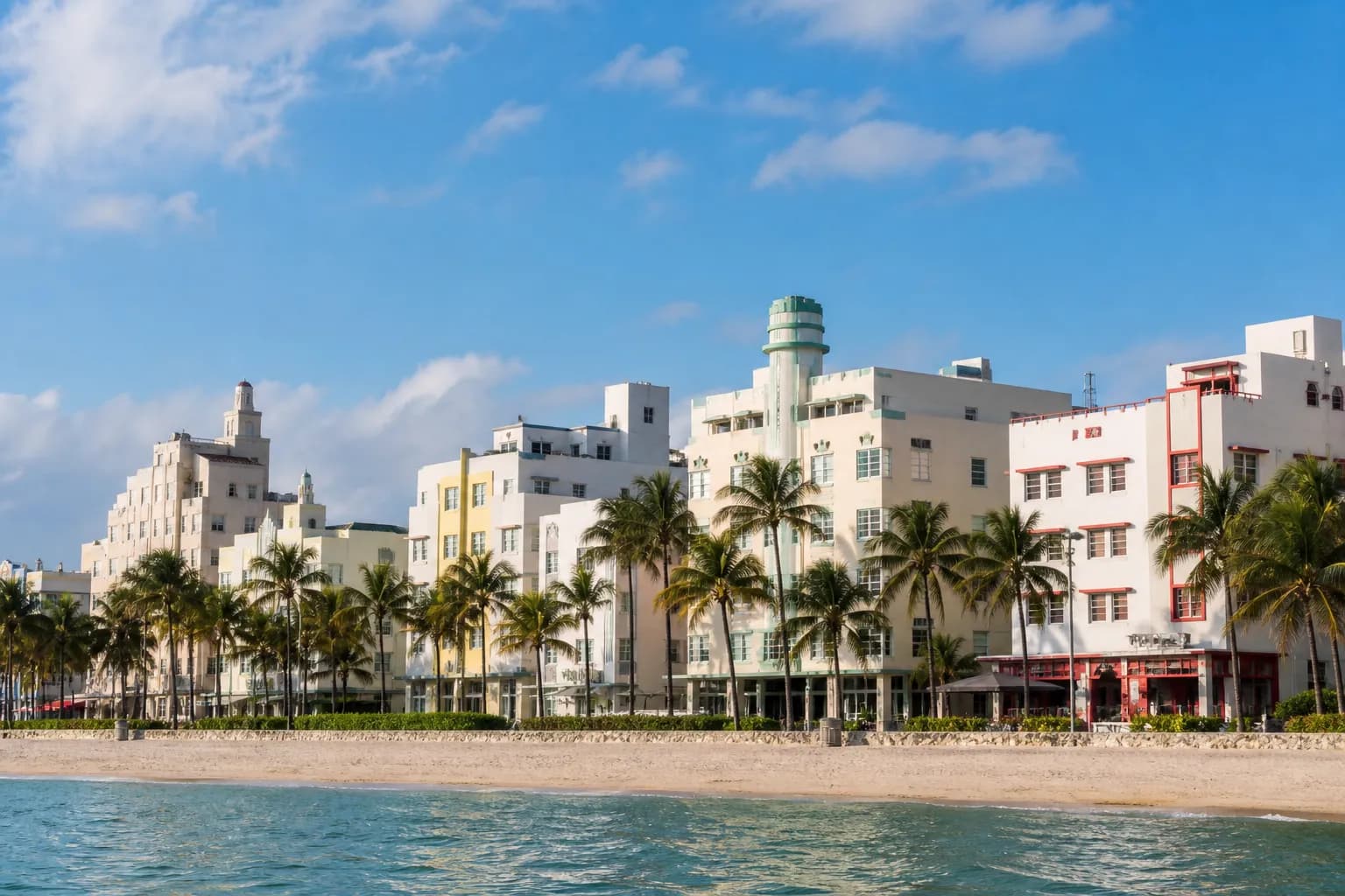 An architectural establishing shot of the Miami Beach shoreline featuring pastel Art Deco buildings, palm trees, and a bright blue sky
