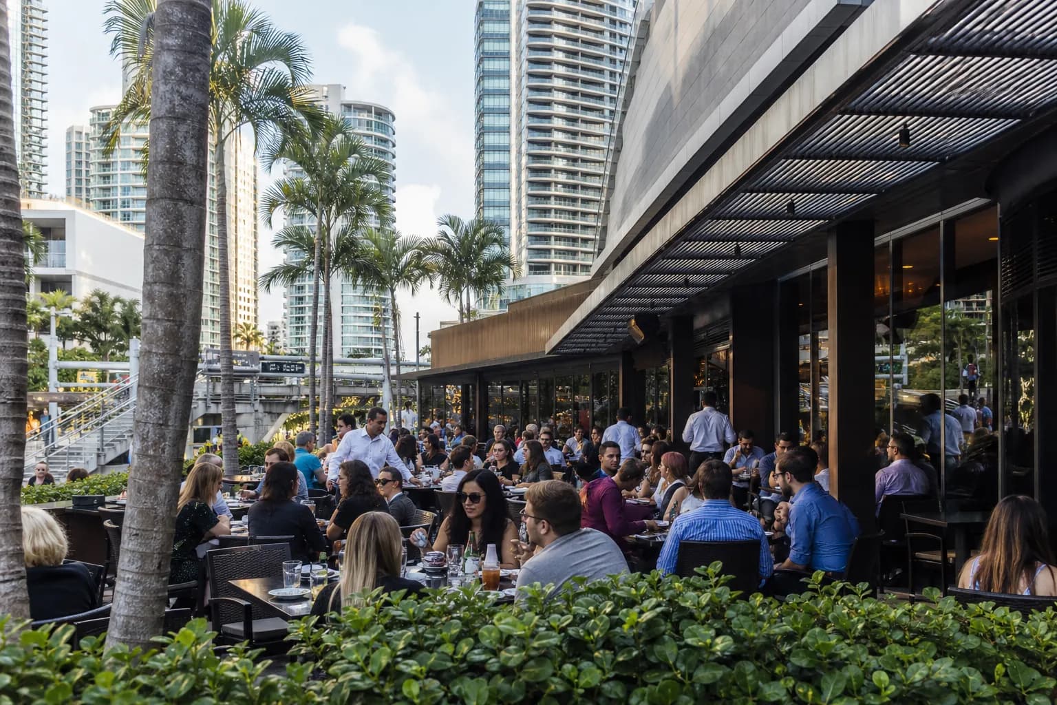 A wide shot of the modern, glass-fronted exterior of the Fogo de Chao restaurant in the bustling Plaza at Brickell in Miami.