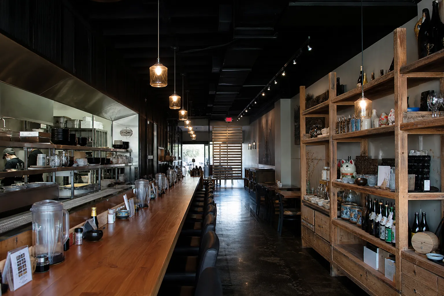 A sleek, long wooden bar counter inside a dimly lit modern Japanese restaurant, with steaming bowls of ramen arranged along the surface.