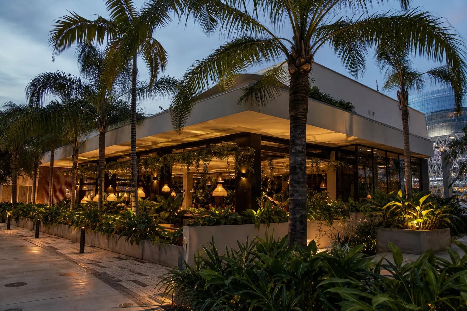 The sleek, modern facade of a contemporary Miami restaurant in the Design District, featuring clean architectural lines, lush tropical foliage, and outdoor seating under warm evening lights.
