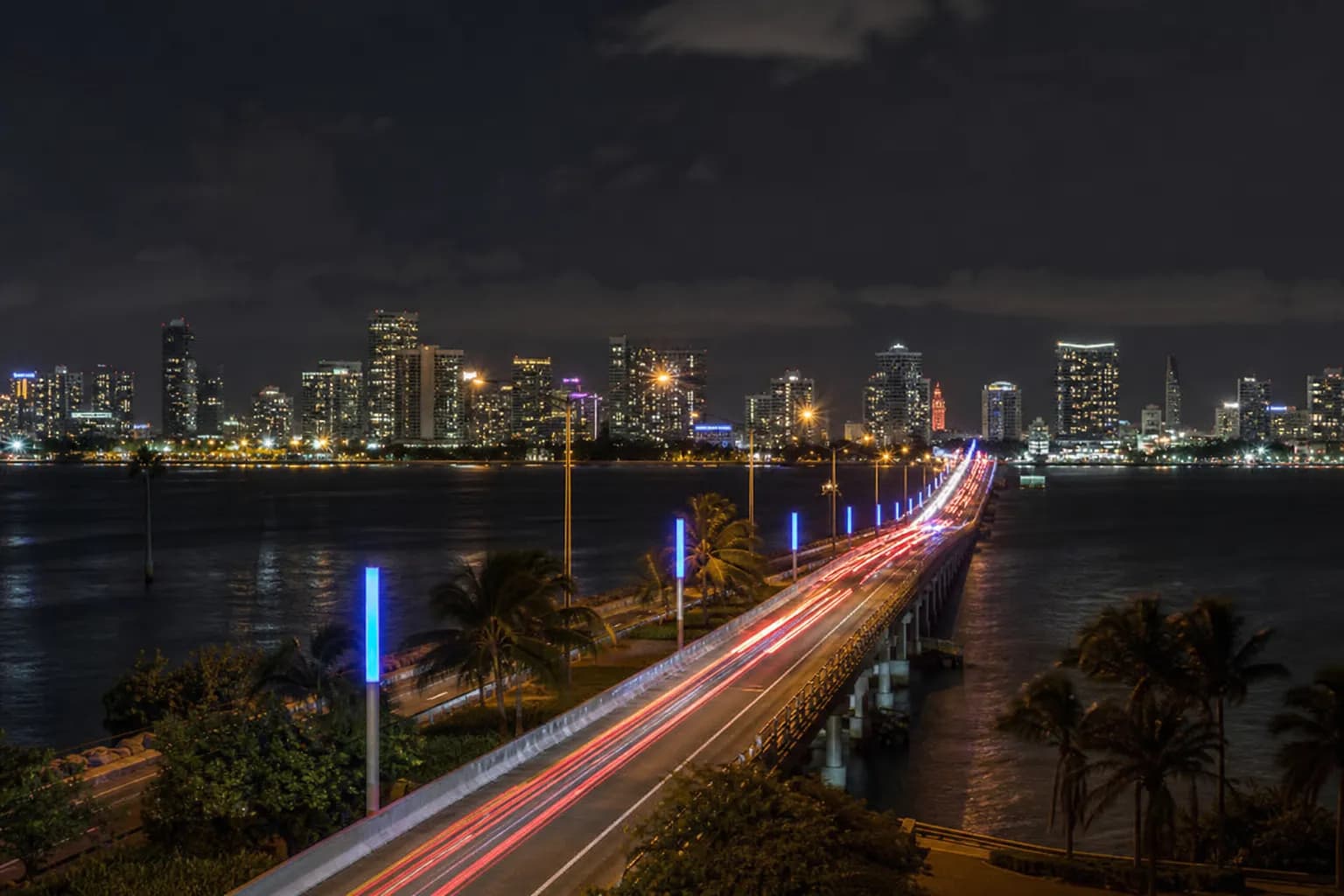 A wide-angle nighttime exterior shot of the neon-lit Julia Tuttle Causeway with car tail lights streaking toward the Miami skyline.