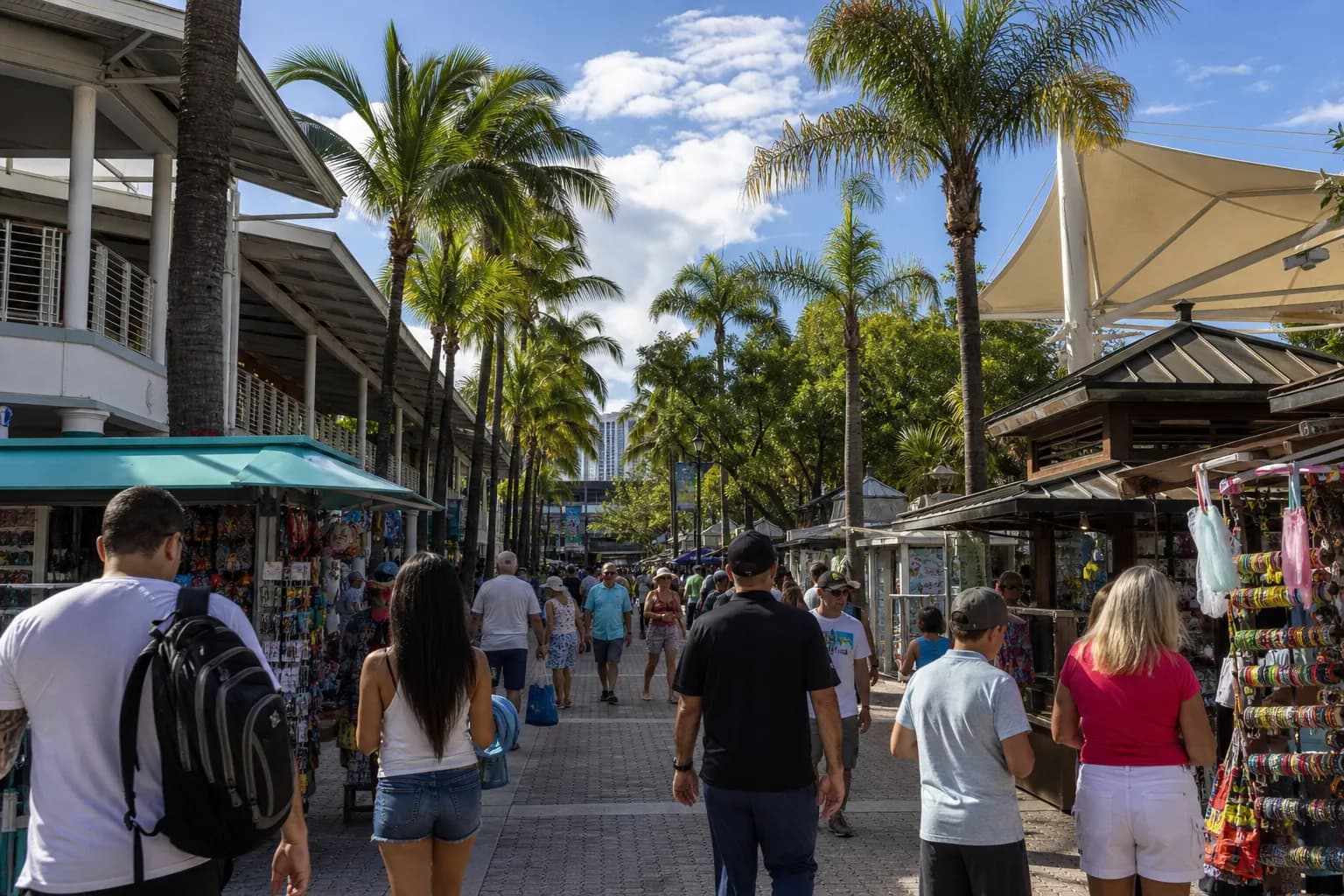 A wide shot looking down a vibrant outdoor market aisle at Bayside Marketplace in Miami, with palm trees and modern storefronts under a bright sky.