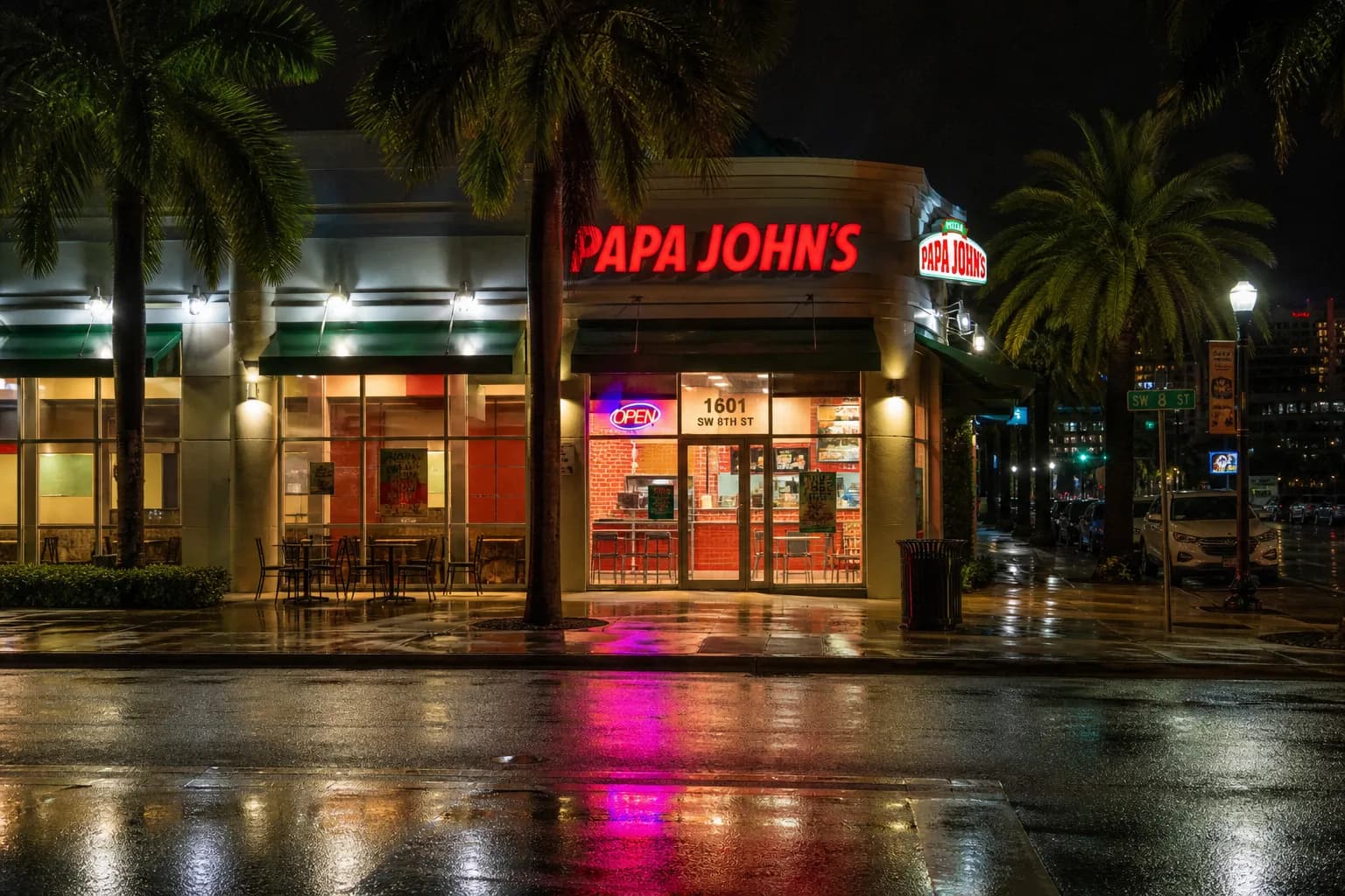 The exterior facade of a Papa Johns pizza store lit up at night along a busy Miami street.