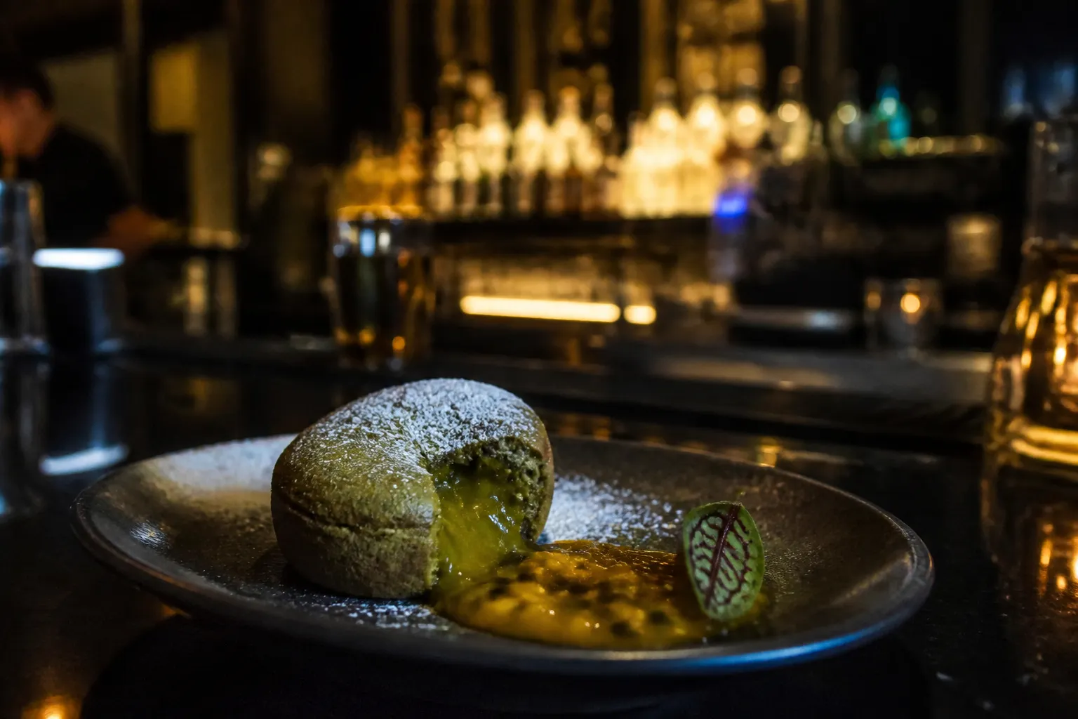 A close-up food still life of a modern Nikkei dessert featuring matcha cake, passionfruit gel, and edible gold leaf on a dark ceramic plate in a moody Brickell restaurant.