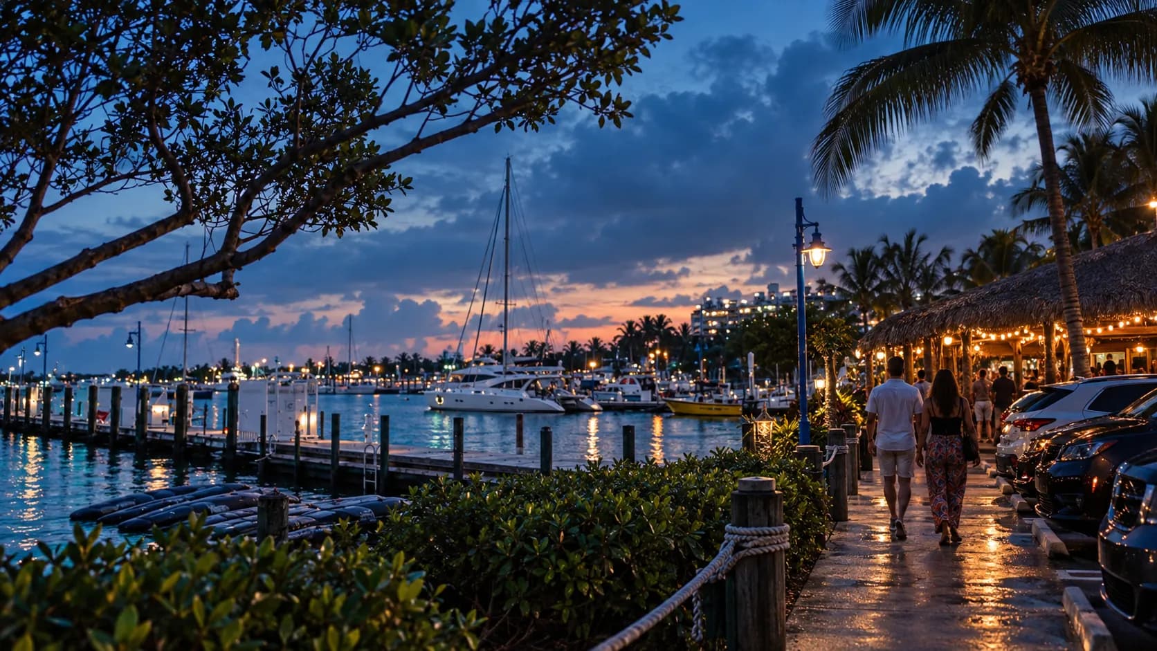 Blue-hour path and parked cars lead to Monty's covered tiki patio, with palms, dock cleats and sailboat masts beyond the darkening bay.