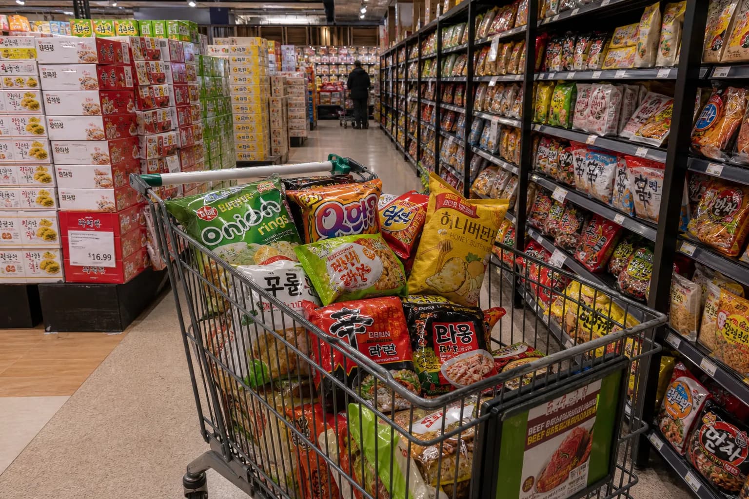 A shopping cart filled with imported Asian snacks and instant ramen in a clean supermarket aisle.