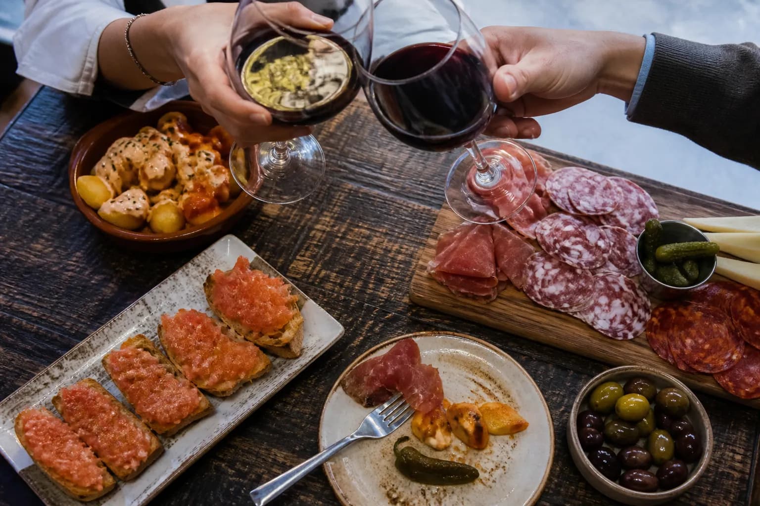 A spread of Spanish tapas including patatas bravas and charcuterie on a rustic table with wine glasses.