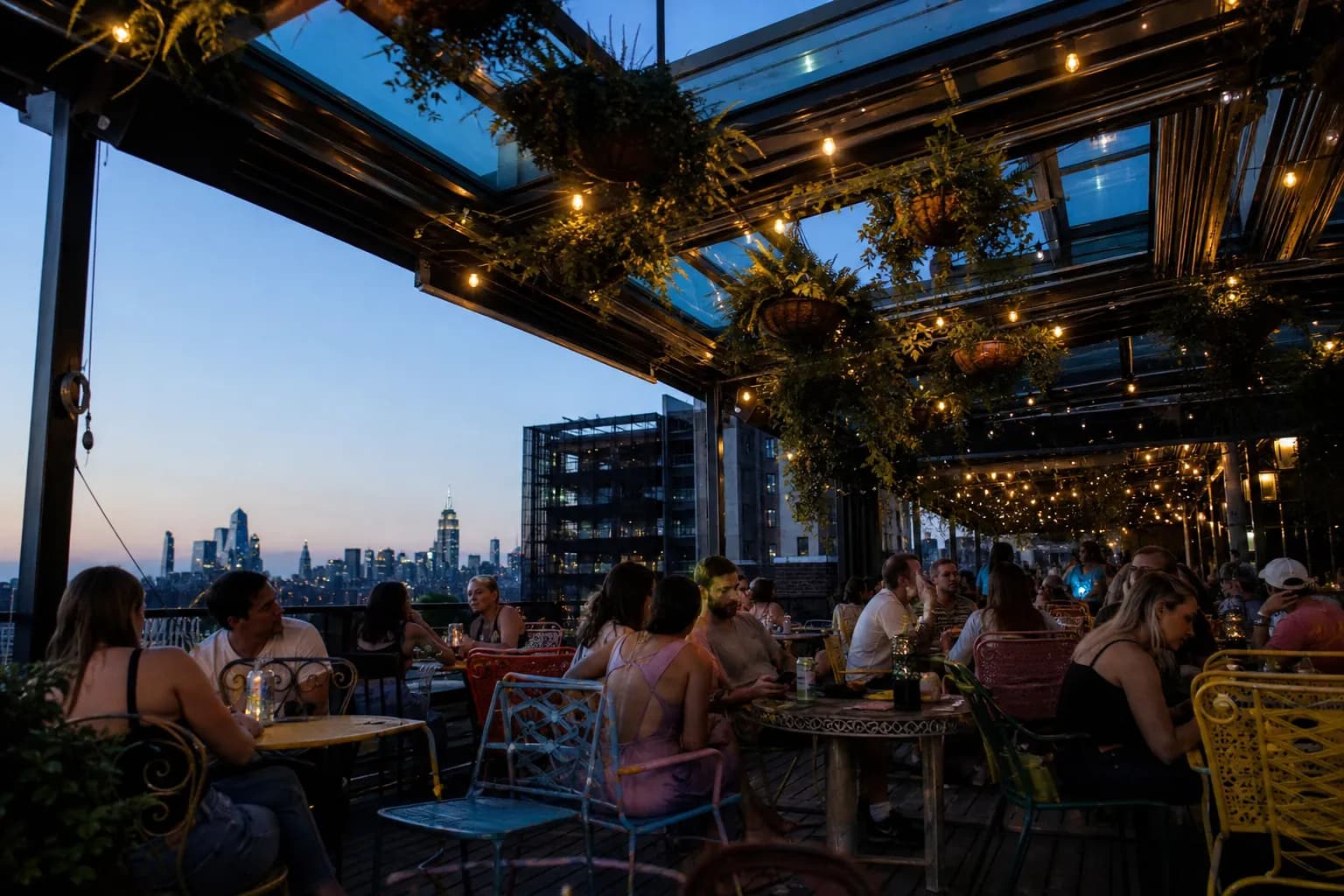 Mismatched patio furniture and hanging greenery under a glass ceiling at a lively East Village rooftop bar during twilight