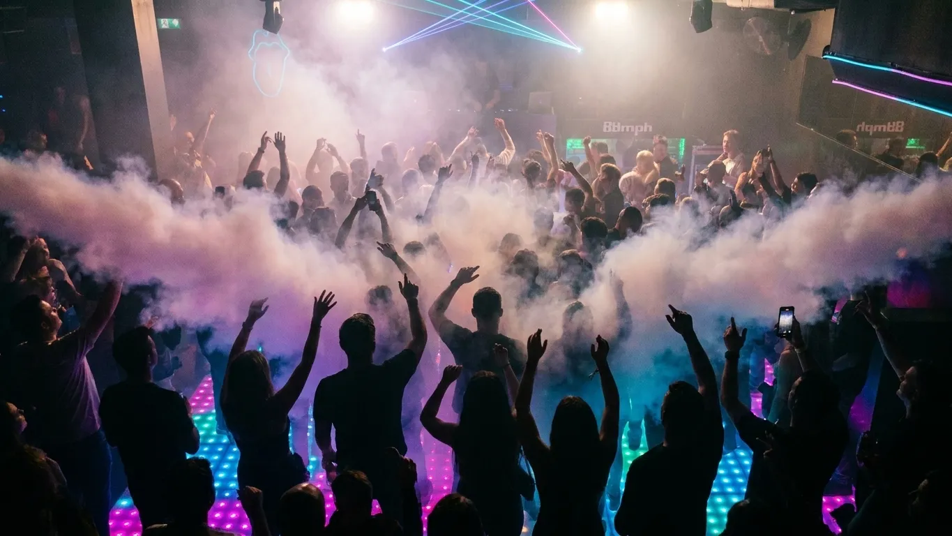 A packed dance floor seen from above with silhouetted shoulders and raised arms in coloured haze and a DJ booth glowing at the far end