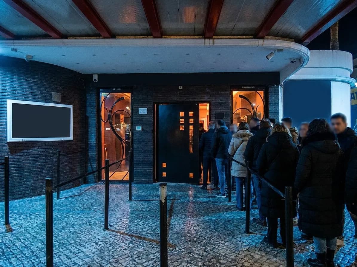 A club entrance at night with a rope and stanchion line along a dark brick facade and a doorway lit from inside